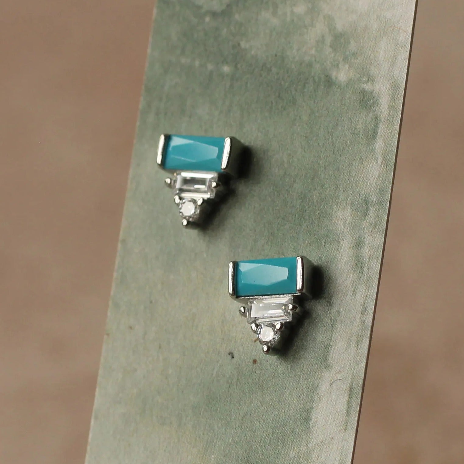 A pair of turquoise-colored earrings with silver accents, displayed on a green card or paper against a blurred background.