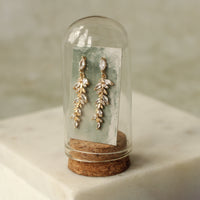 A pair of elegant gold earrings with clear gemstones, displayed in a glass case with a cork base.