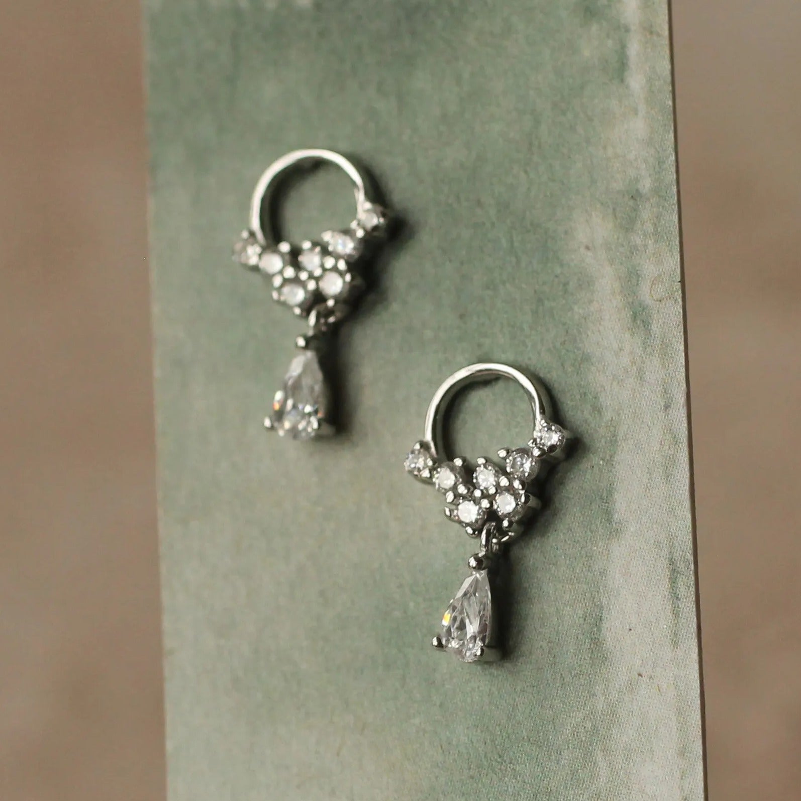 A pair of elegant silver earrings with small, clear stones hanging from the center, displayed on a green card.