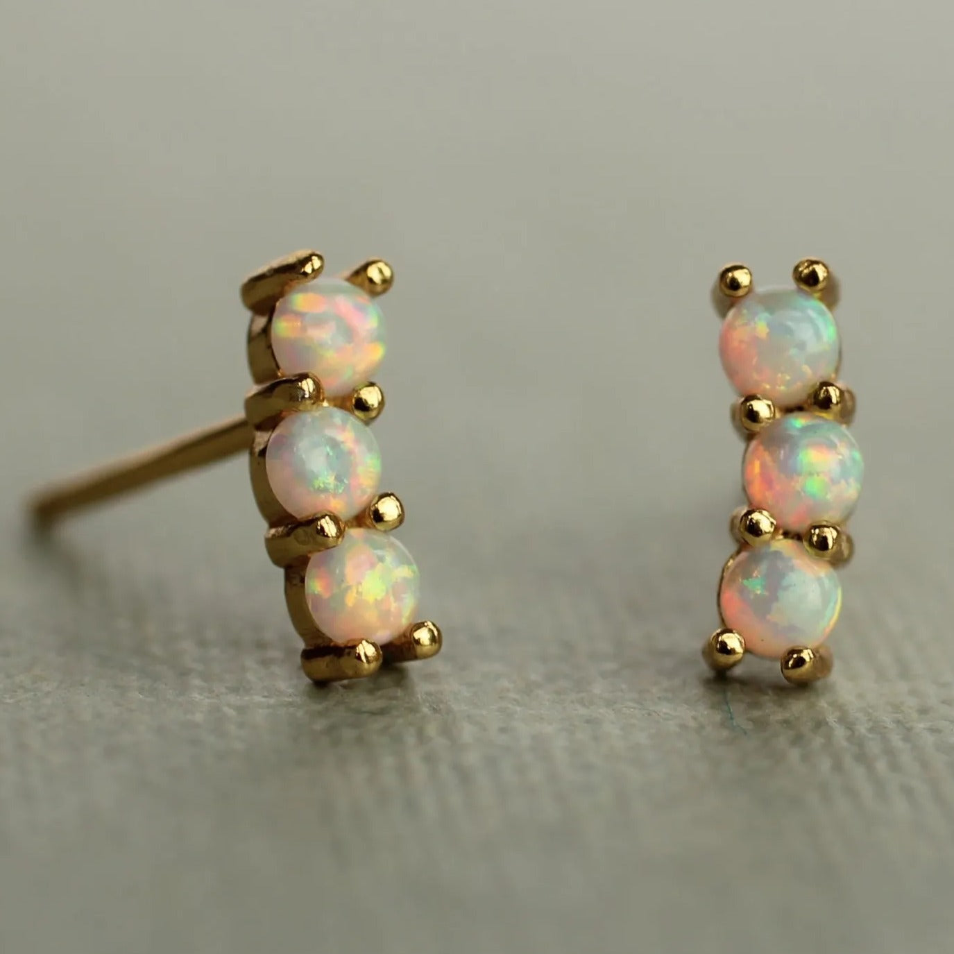 A pair of gold earrings with three opal stones set in a row, resting on a textured surface.