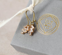 A pair of gold earrings with clear gemstone accents, hanging from a white ribbon, and a gray box with a gold embossed logo.