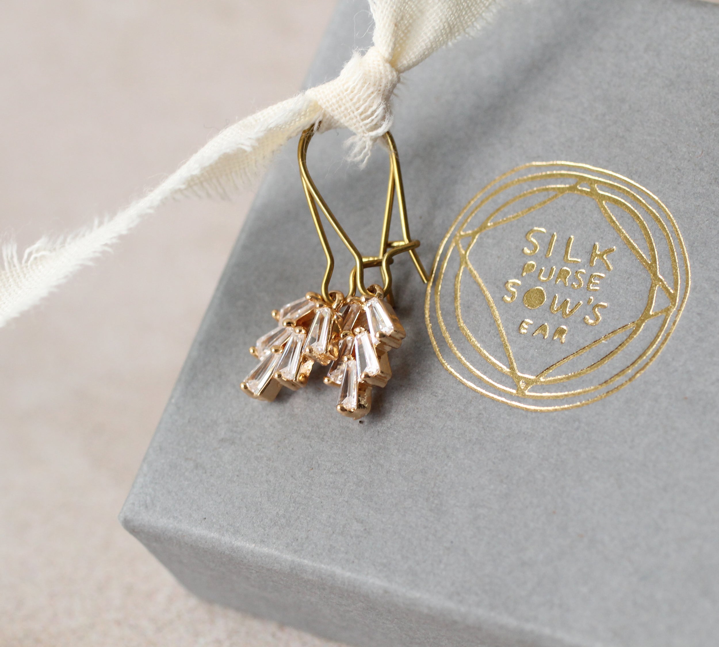 A pair of gold earrings with clear gemstone accents, hanging from a white ribbon, and a gray box with a gold embossed logo.