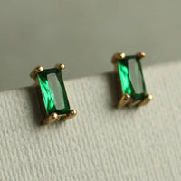 A pair of gold earrings with green gemstones, specifically emeralds, are displayed on a white surface.