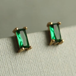 A pair of gold earrings with green gemstones, specifically emeralds, are displayed on a white surface.