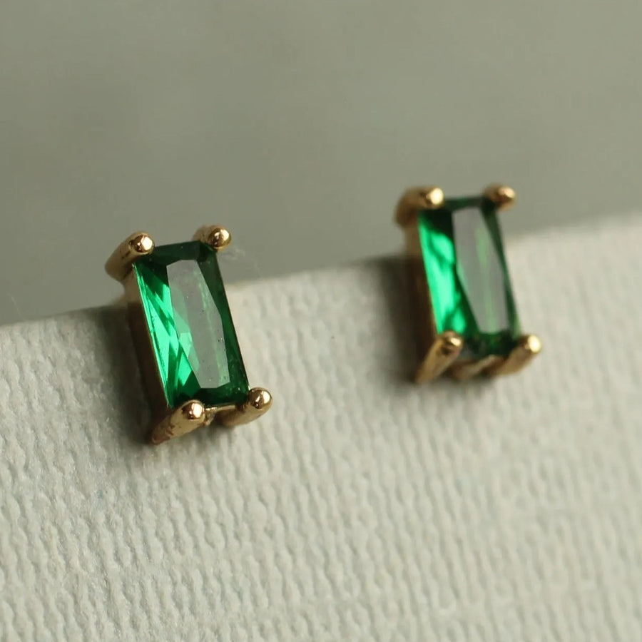 A pair of gold earrings with green gemstones, specifically emeralds, are displayed on a white surface.