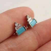 A close-up image of a person's hand holding a pair of small, turquoise-colored stud earrings with silver and clear gemstone accents.