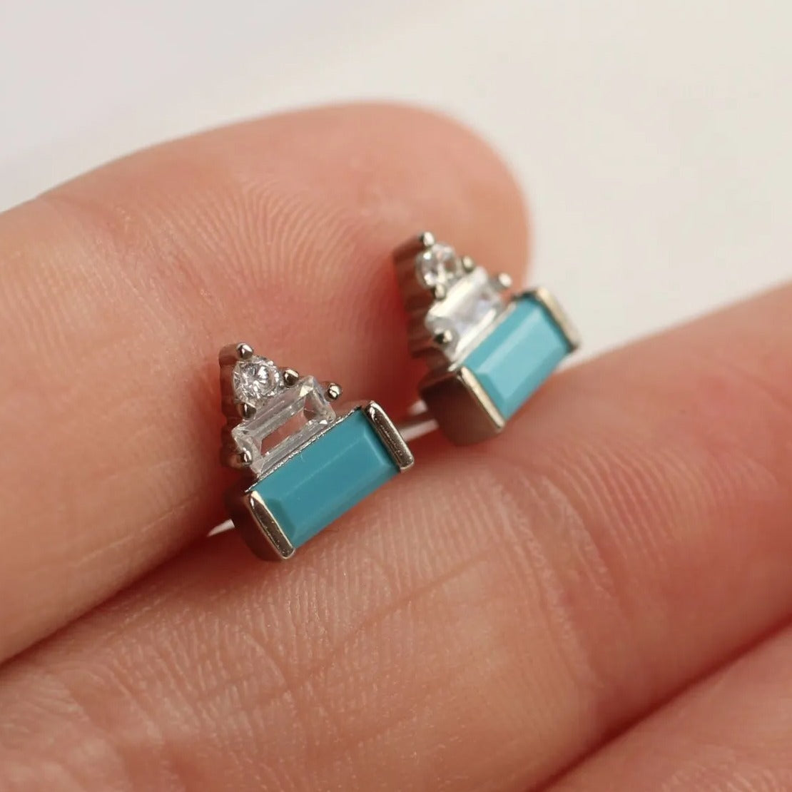 A close-up image of a person's hand holding a pair of small, turquoise-colored stud earrings with silver and clear gemstone accents.