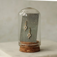 A pair of gold earrings with clear gemstones is displayed in a glass case with a cork base.