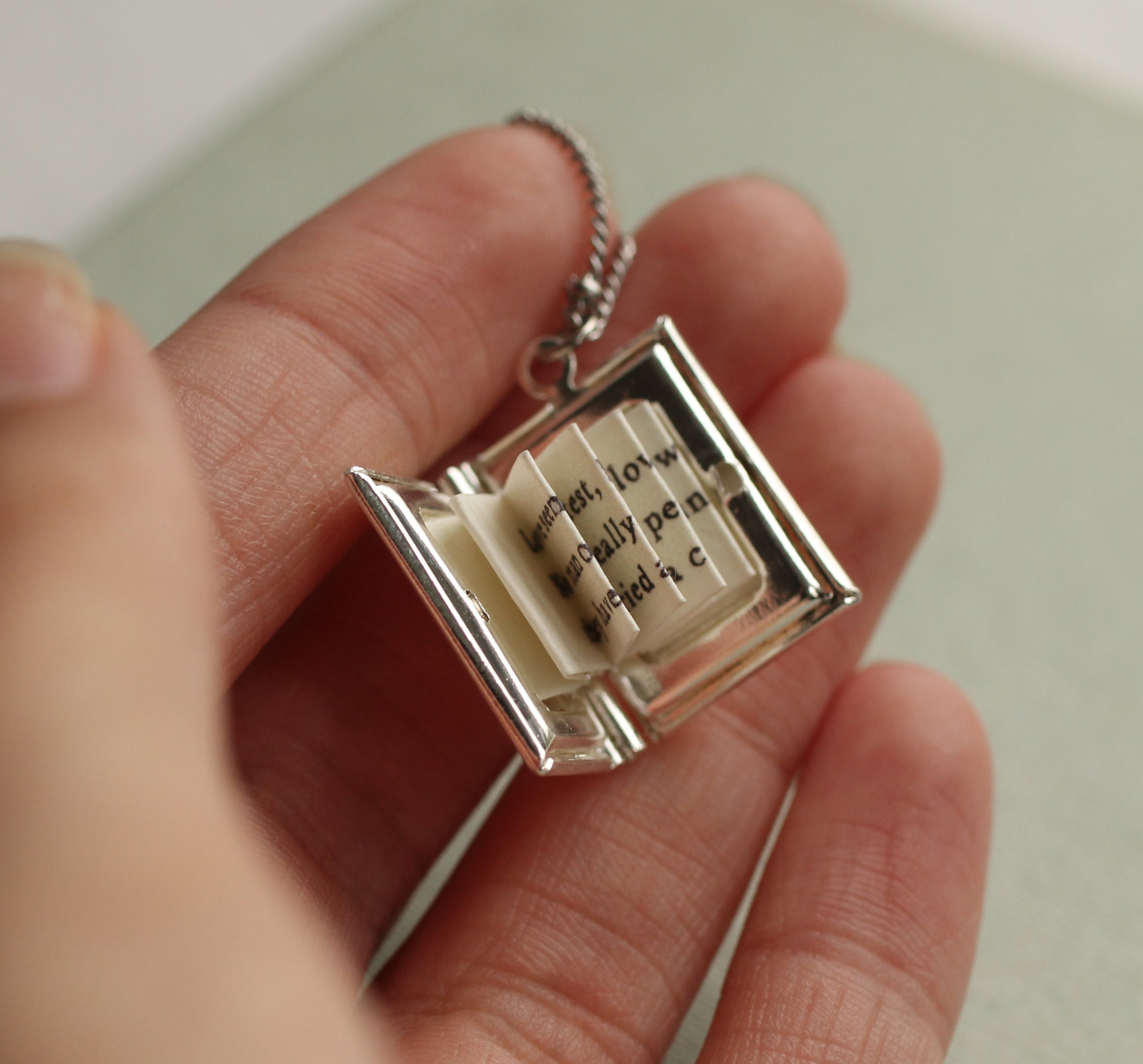A person's hand holding a small silver-colored charm that appears to be an open book or a miniature book-like object.