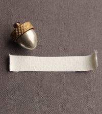 a small, shiny acorn-shaped object with a golden top, placed next to a piece of white paper on a textured gray background.