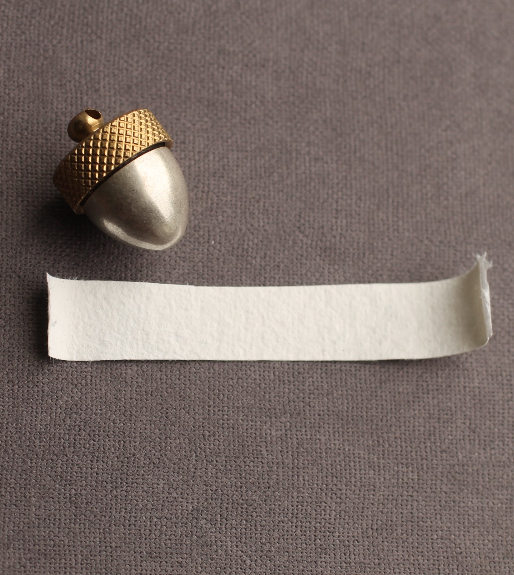 a small, shiny acorn-shaped object with a golden top, placed next to a piece of white paper on a textured gray background.