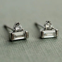 A pair of silver earrings with clear gemstones, resting on a textured surface.