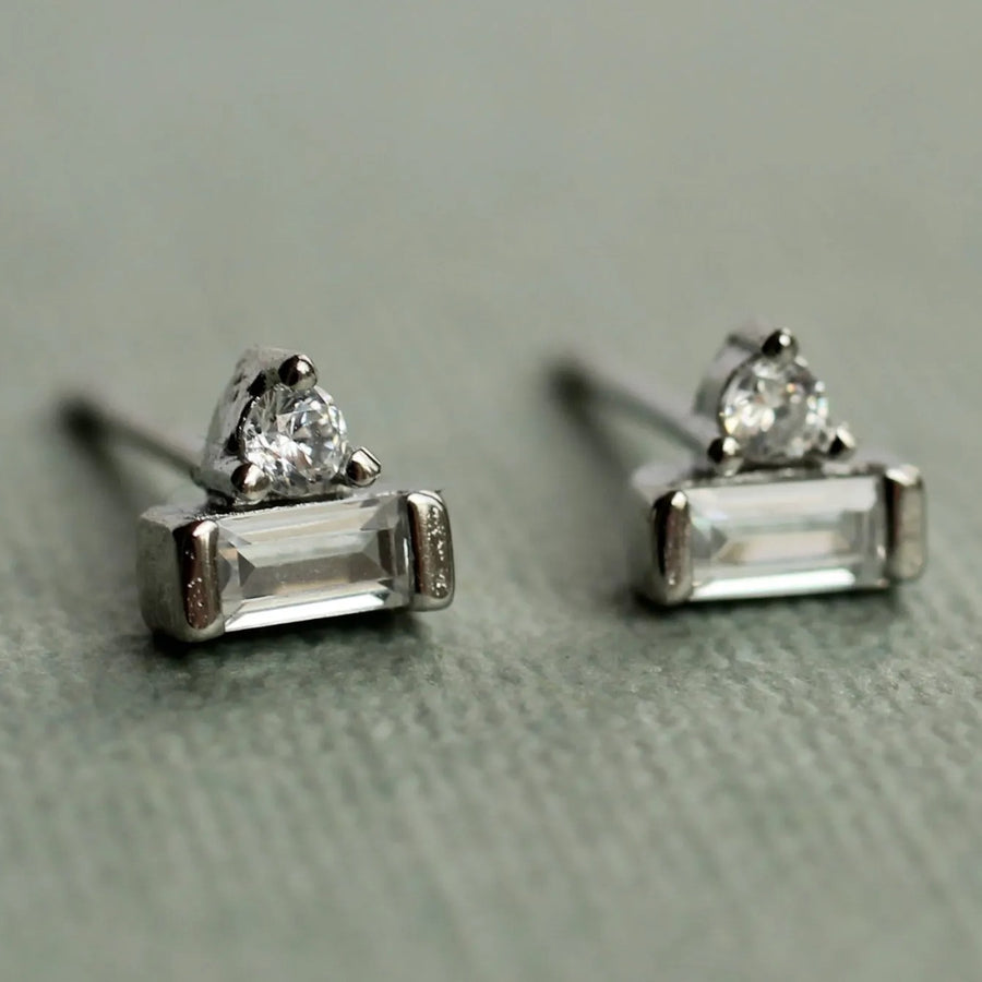 A pair of silver earrings with clear gemstones, resting on a textured surface.