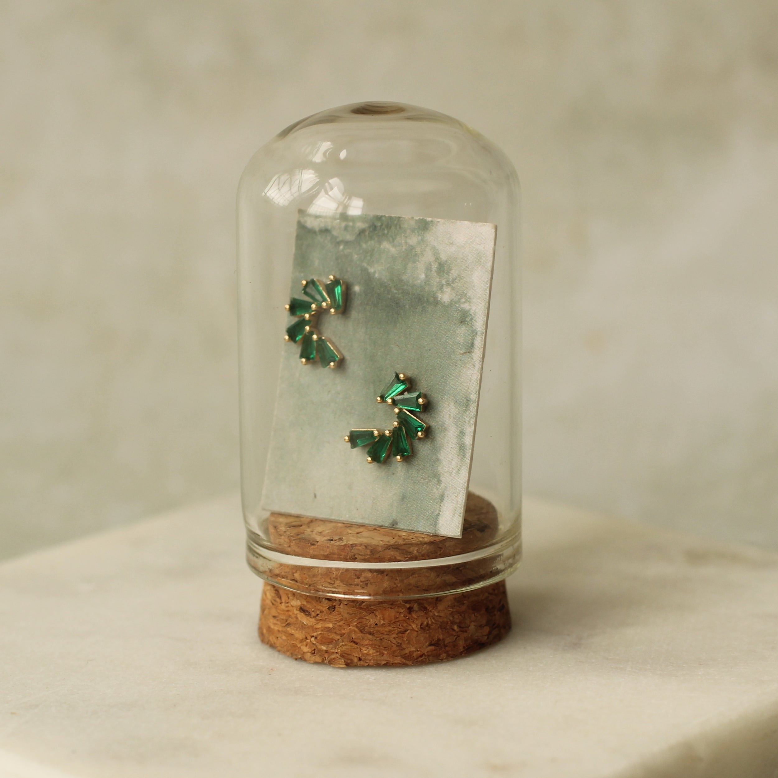 A pair of green earrings is displayed in a glass dome, with a cork base, against a white background.