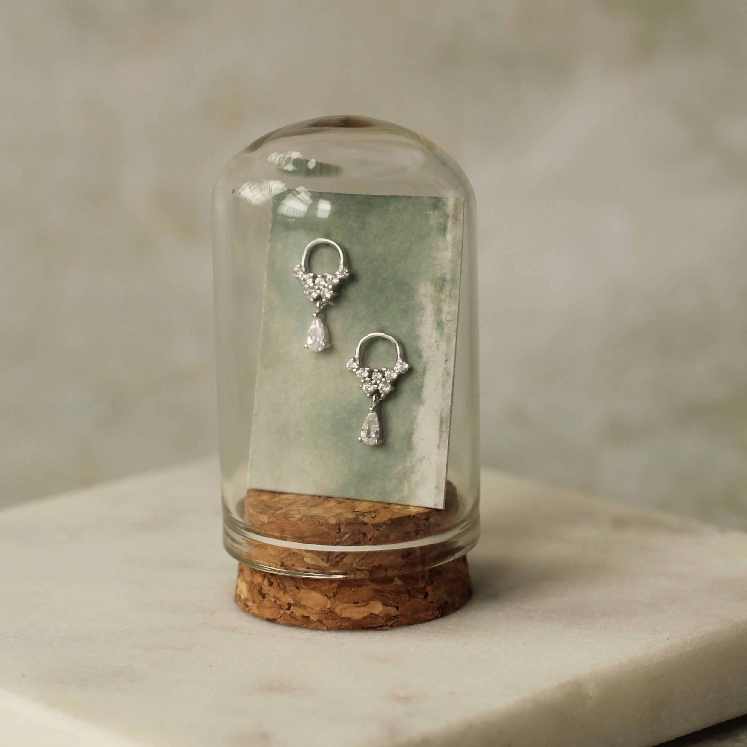 A pair of silver earrings with dangling designs, displayed in a glass dome on a cork base.