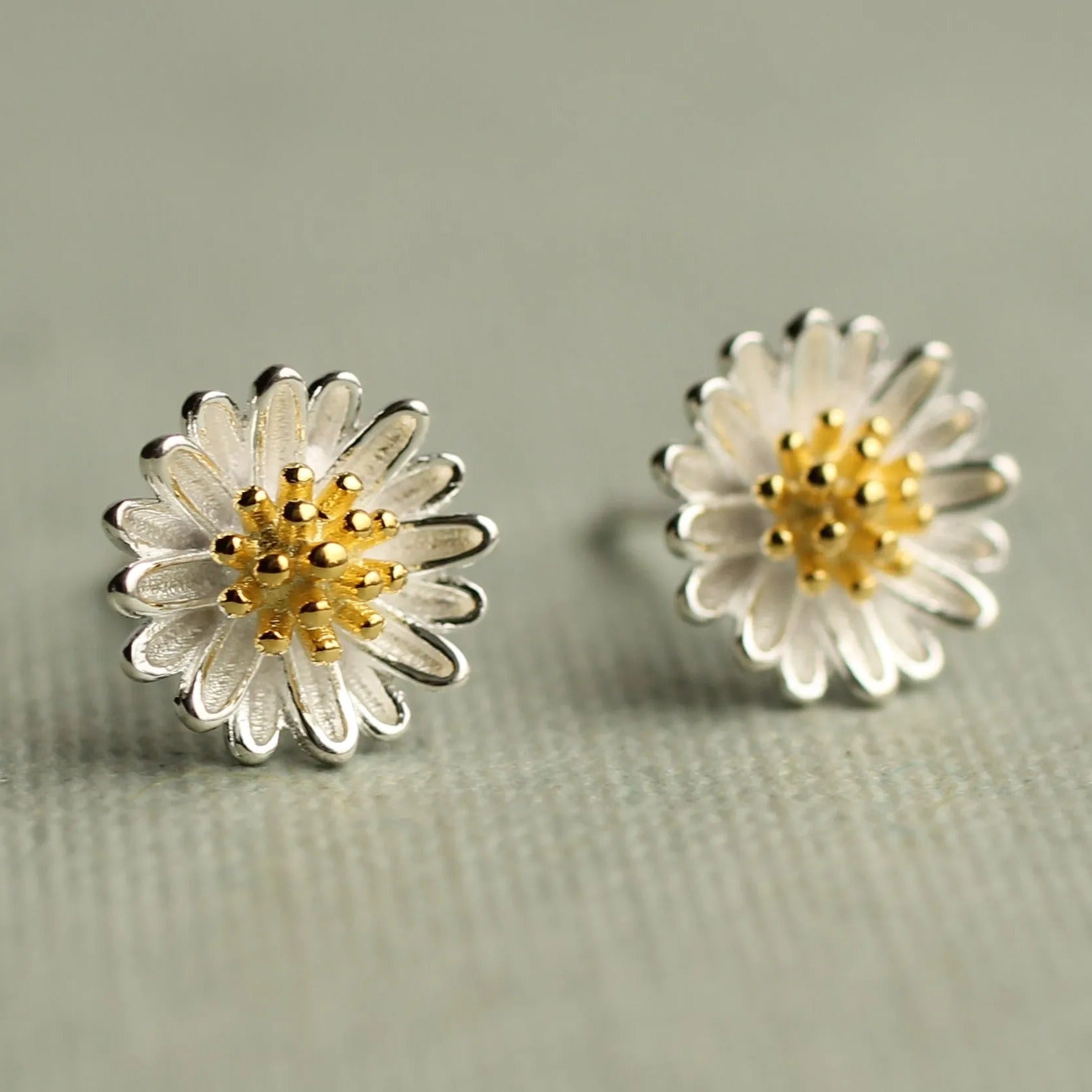 a pair of silver earrings with white and gold flower-shaped designs, placed on a textured surface.