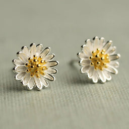 a pair of silver earrings with white and gold flower-shaped designs, placed on a textured surface.