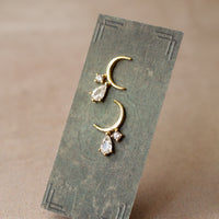 A pair of gold earrings with small clear stones hanging from them, displayed on a gray card.