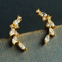 A pair of gold earrings with clear gemstones, such as diamonds, set in a decorative design, resting on a textured fabric surface.