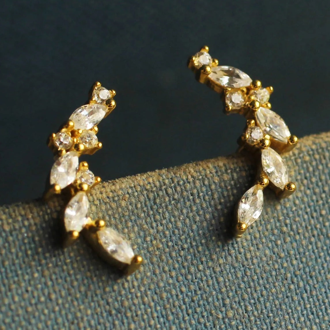 A pair of gold earrings with clear gemstones, such as diamonds, set in a decorative design, resting on a textured fabric surface.